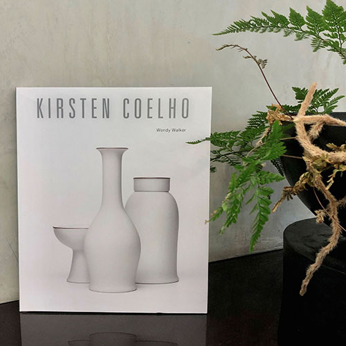 Image of the Kirsten Coelho SALA Monograph book sitting on a table next to a fern. The book is white with three white ceramic vessels on the cover.jpg