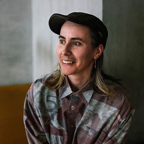 Portrait of Amos Gebhardt, a Caucasian person with shoulder length blonde hair, wearing a black cap, thin, dangly earring and patterned shirt.jpg
