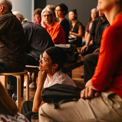 Photo of Vernon Ah Kee sitting in front of one of his text based works. Vernon is an Aboriginal man with short dark hair wearing a navy suit jacket over a black shirt and dark pants.jpg