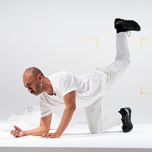 Photo of Vernon Ah Kee sitting in front of one of his text based works. Vernon is an Aboriginal man with short dark hair wearing a navy suit jacket over a black shirt and dark pants.jpg