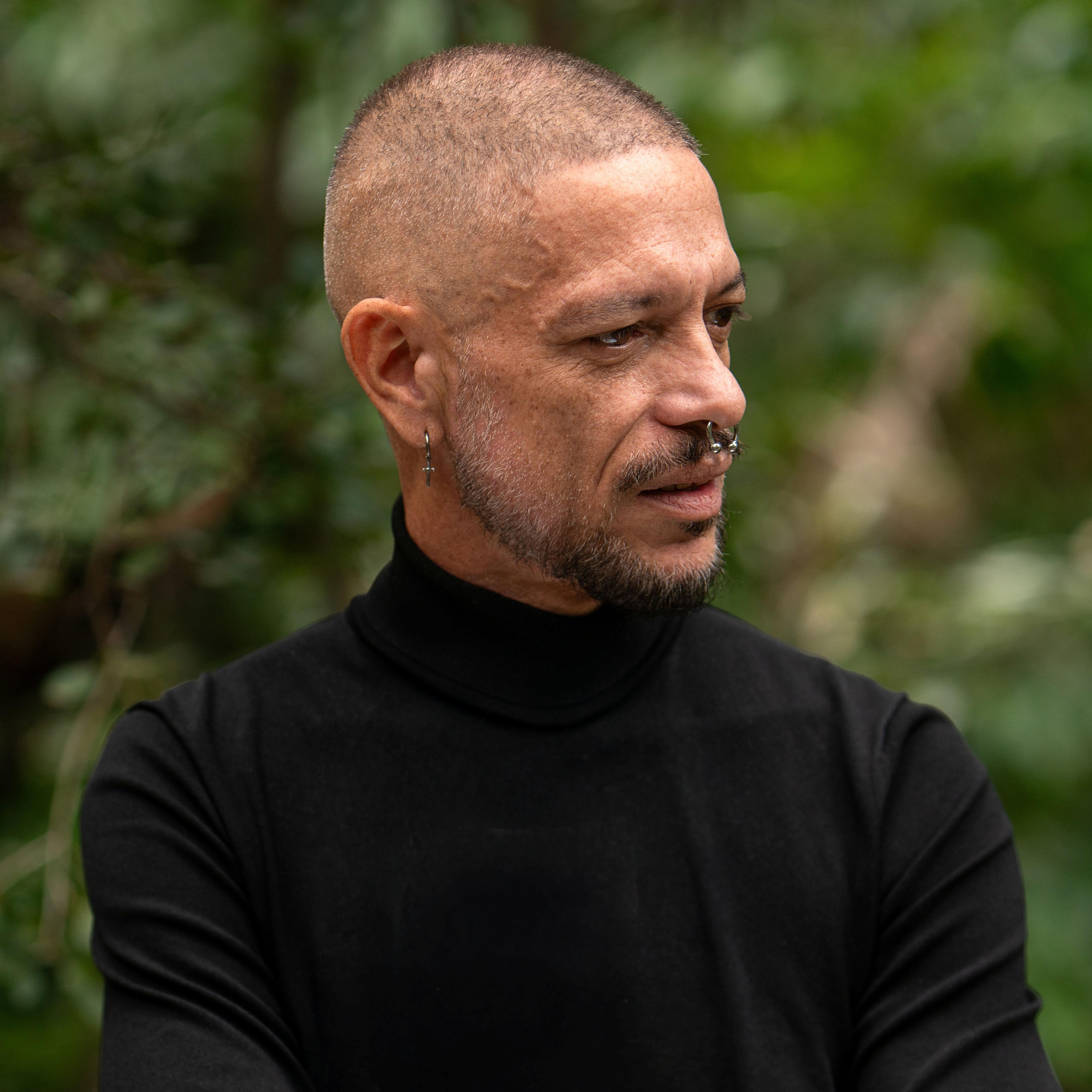 Photo of Vernon Ah Kee sitting in front of one of his text based works. Vernon is an Aboriginal man with short dark hair wearing a navy suit jacket over a black shirt and dark pants.jpg