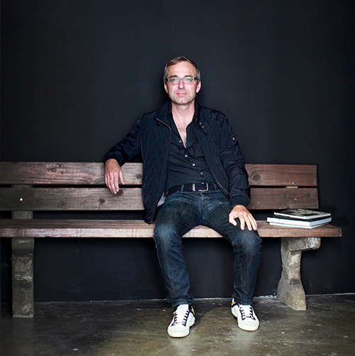 Portrait of Belgian artist David Claerbout sitting on a wooden bench wearing light coloured glasses, a navy jacket and shirt, indigo jeans and white sneakers.jpg