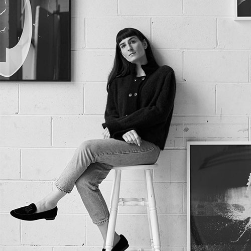 Black and white portrait of Zoe Croggon sitting on a stool, leaning against a brick wall. Zoe is a Caucasian female with long dark hair wearing a dark top and jacket, pale jeans and dark loafers.jpg