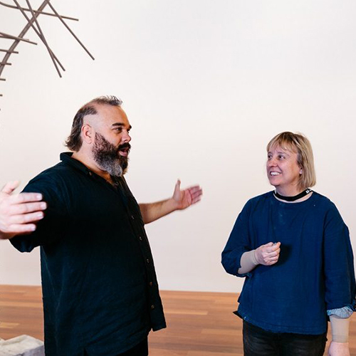 Monte Masi, a Caucasian male with dark hair and beard wearing a black shirt and pants, stands with his arms outstretched as he speaks to Louise Haselton, a Caucasian female with light shoulder length hair wearing a blue top and dark trousers..jpg