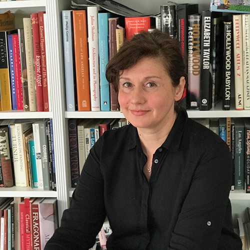 Portrait of Anne Wallace sitting in front of a bookshelf full of books. Anne is a Caucasian woman with dark brown curly hair, pulled back into a tail, wearing a black shirt.jpg