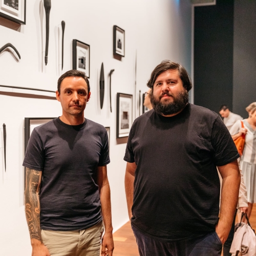 Portrait of James Tylor and Dominic Guerrera standing in front of Jame's installation of photos and objects hung on the gallery wall at Samstag. James is an Aboriginal man with short dark hair and tattoos down his right arm, wearing a dark t-shirt and beige pants. Dominic is an Aboriginal man with ear-length, dark-brown hair and beard, wearing a dark t-shirt and navy pants.jpg