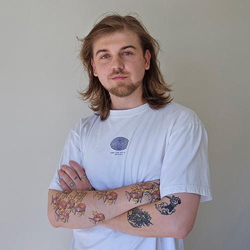 Portrait of Ash Tower, a Caucasian male with shoulder length, light brown hair and tattoos on both arms wearing a white t-shirt with blue logo.jpg